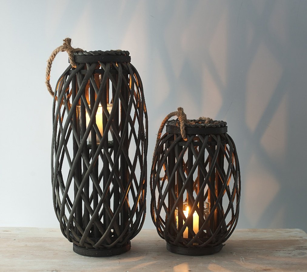 A dark toned woven wicker lantern with a chunky rope handle accent and glass insert 