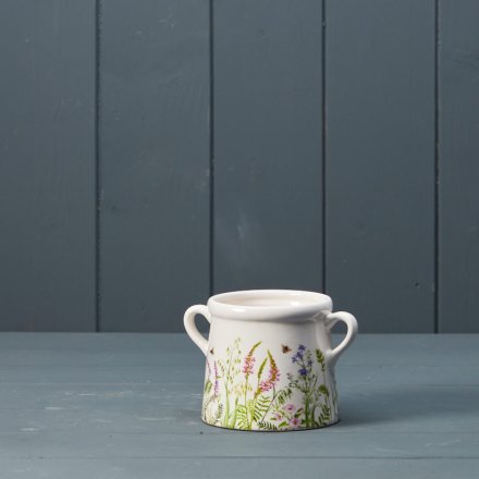 A dainty white pot with two handles featuring a charming meadow design. 
