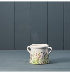 A dainty white pot with two handles featuring a charming meadow design. 